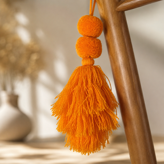 Orange pom-pom tassel hanging decoration with two pom-poms and layered orange yarn tassels hanging on a wooden chair.