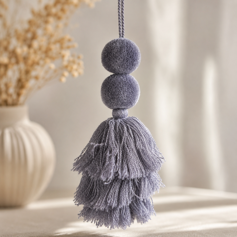 Grey pom-pom tassel hanging decoration with two pom-poms and layered grey yarn tassels hanging on a wooden chair.