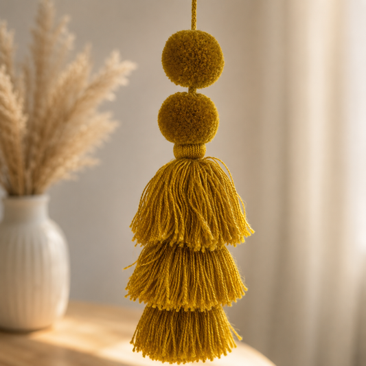 A mustard yellow decorative tassel ornament with three pom-poms and layered fringe, hanging in front of a softly blurred indoor background with a white vase and dried grasses.