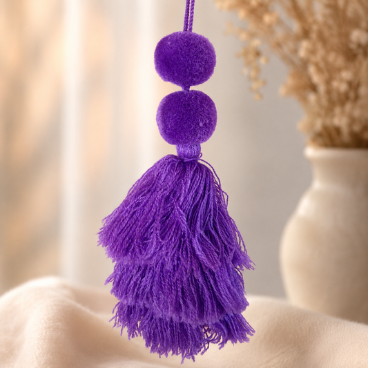 A purple velvet tassel ornament with two round pom-poms and a large layered tassel, hanging against a softly blurred beige background with dried flowers in a vase.