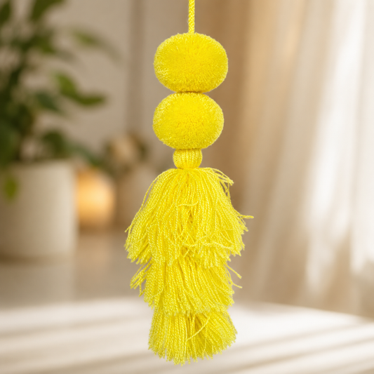 A bright yellow decorative hanging ornament with two round pom-poms and a large tassel, shown against a softly blurred indoor background with curtains and a plant.