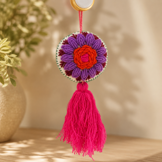 Handmade embroidered floral ornament with purple petals, orange center, and pink tassel on yellow background