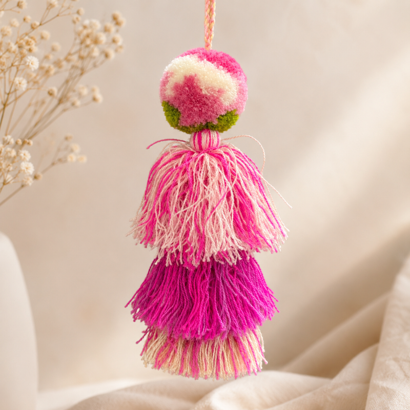 Blush pink pom pom tassel bag charm with layered pink tassels attached to a beige handbag