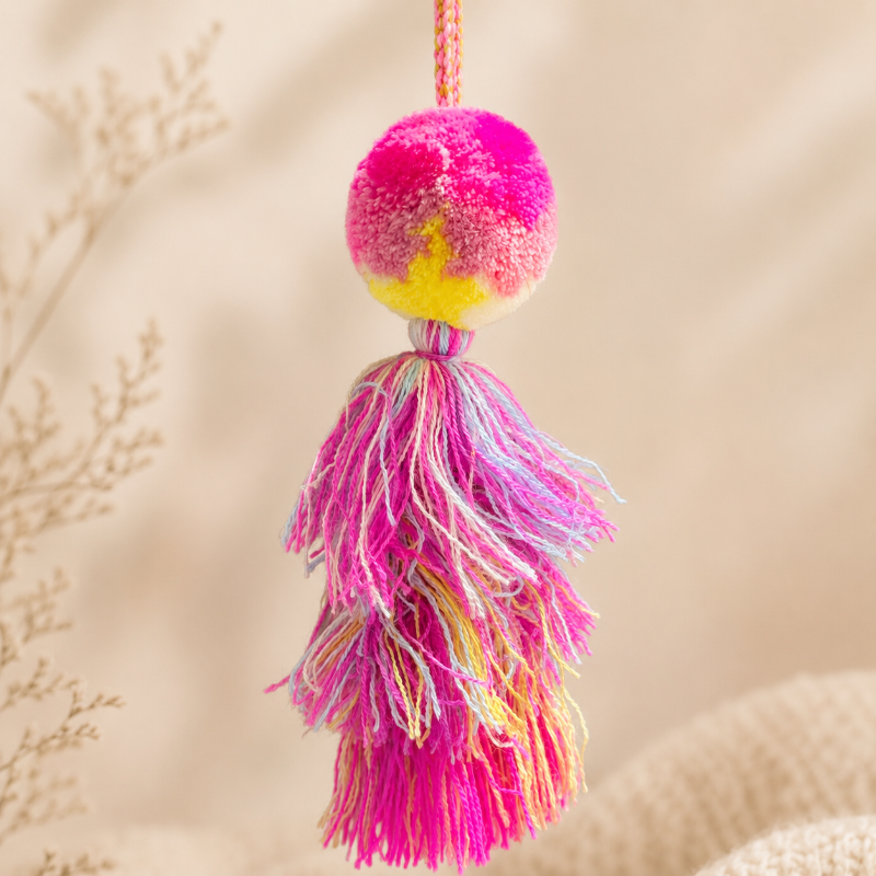 Hot pink pom pom tassel bag charm with layered multicolor tassels attached to a beige handbag