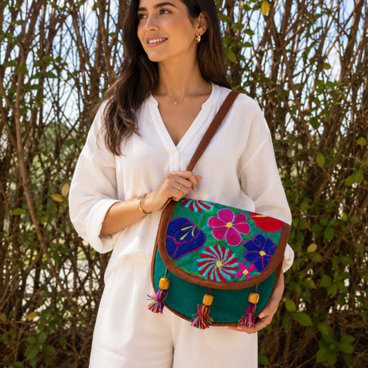 A woman in a white outfit carries a colorful embroidered shoulder bag with a green base, pink and blue floral patterns, brown trim, and tassel details, set against a background of leafy green bushes.