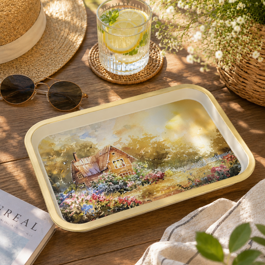A rectangular metal tray with rounded corners featuring a colorful watercolor painting of a cottage in a flower field, styled on a wooden table with a straw hat, sunglasses, a glass of lemonade, and flowers.