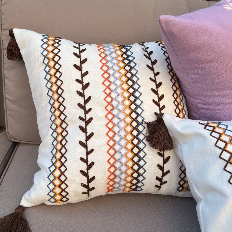 Square boho cushion cover with multi-color embroidered geometric and leaf pattern, finished with brown tassels on all four corners, 43x43 cm