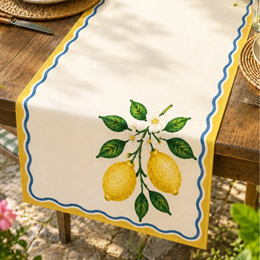 An off-white table runner with a yellow scalloped border, blue wavy inner border, and a green and yellow lemon illustration, laid on a wooden outdoor table.