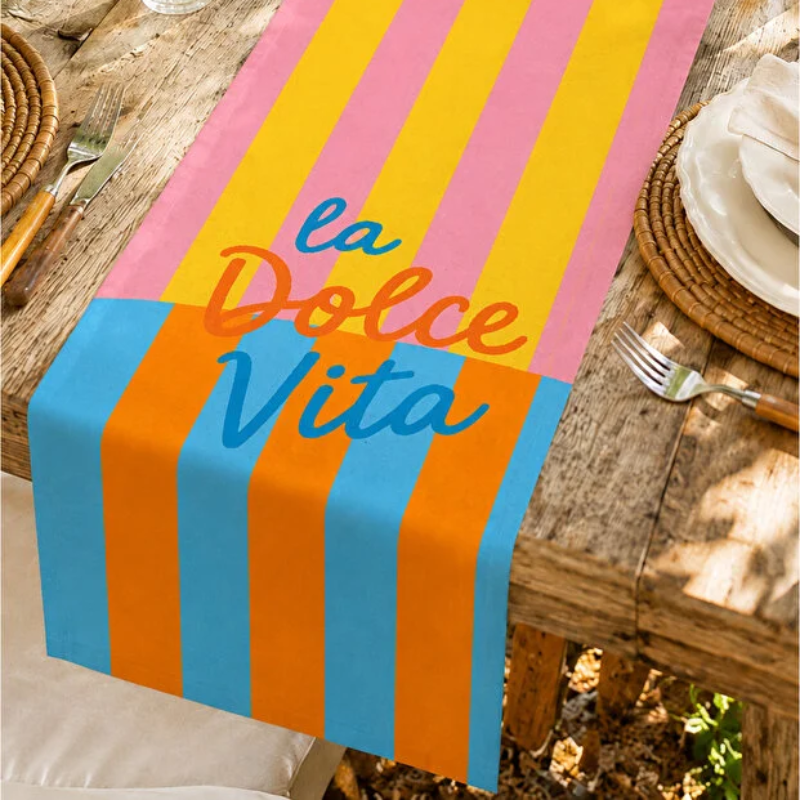 A colorful table runner laid on a rustic wooden dining table, featuring vertical pink, yellow, blue, and orange stripes with the words 'la Dolce Vita' in stylized script.