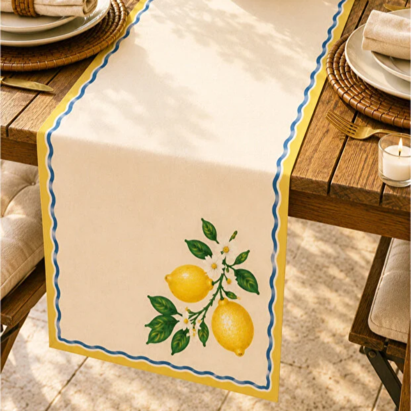 A cream table runner with a yellow border, blue scalloped edge, and embroidered lemon motif, laid on a wooden dining table with plates and a candle.