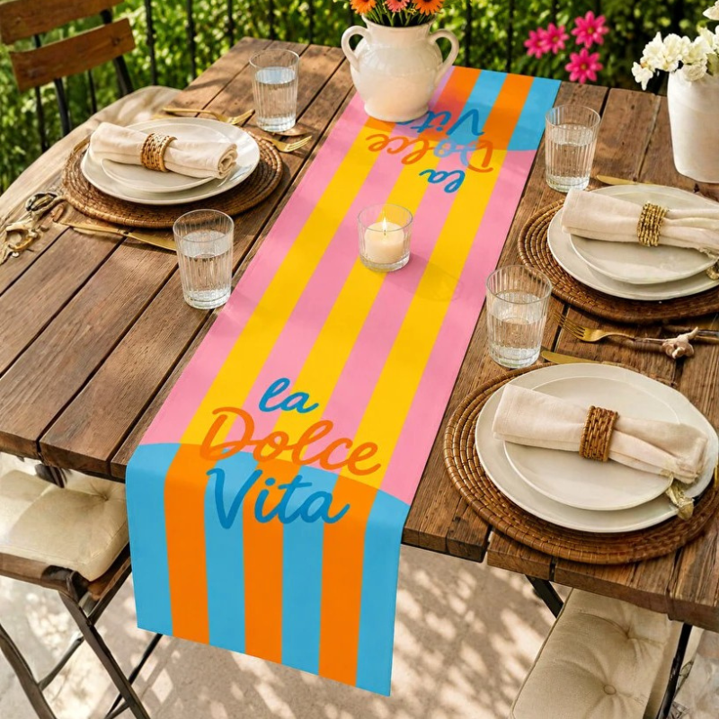 A colorful table runner with vertical stripes in pink, yellow, orange, and blue, printed with the text 'la Dolce Vita' in a script font. The runner is styled on a wooden outdoor dining table with plates, napkins, glasses, and a candle.