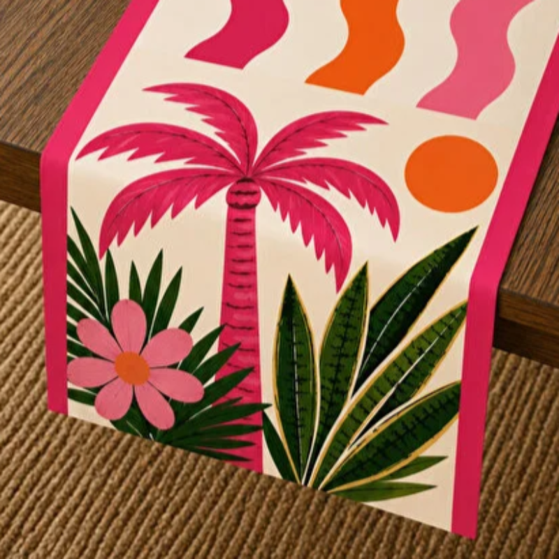 A tropical-themed table runner with a cream background, featuring a pink palm tree, green leaves, a pink flower, and abstract wavy shapes in pink, orange, and green, draped over a wooden table.