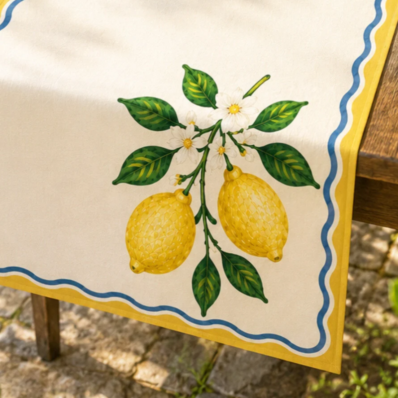 A cream-colored table runner with a scalloped yellow and blue border, featuring a detailed illustration of lemons, green leaves, and white flowers, draped over a wooden table outdoors.
