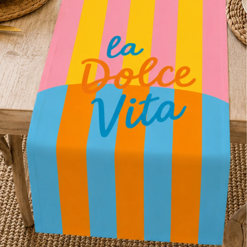A colorful table runner draped over a wooden table, featuring vertical stripes in pink, yellow, blue, and orange with the text 'la Dolce Vita' in stylized lettering.