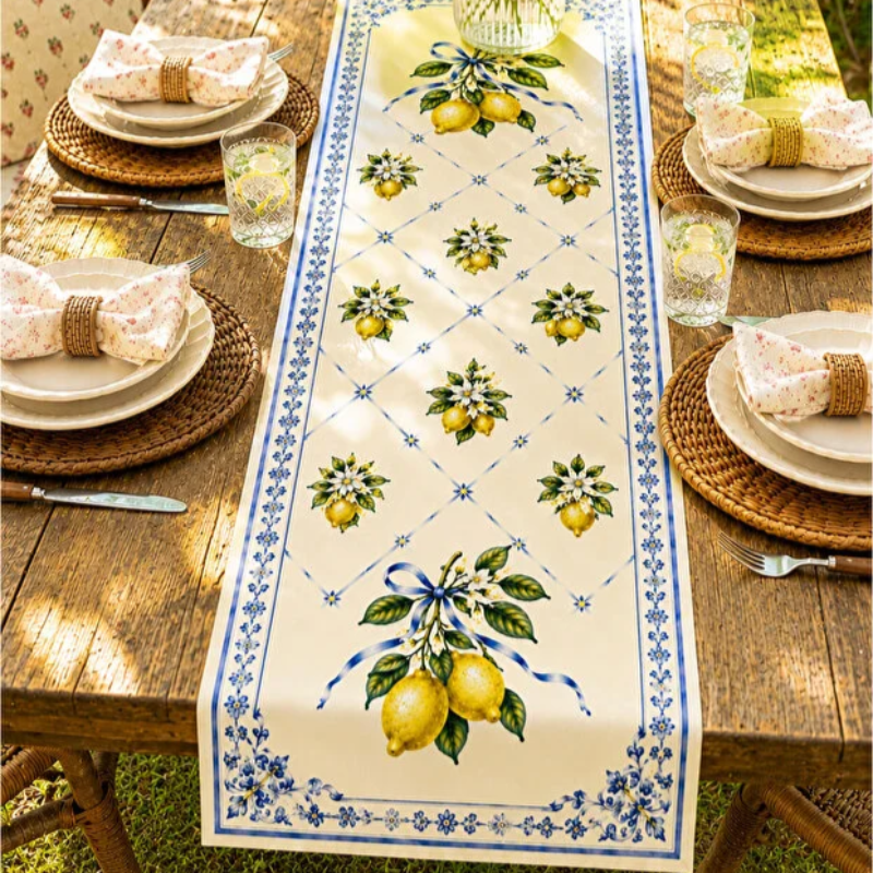 A rectangular table runner laid on a wooden outdoor table, featuring a cream background with blue floral borders and repeated lemon and green leaf motifs. Plates, napkins, glasses, and woven placemats are arranged around it.