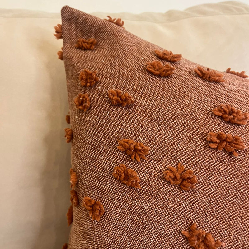 Rust textured square throw pillow with raised pom pom details placed on a beige sofa