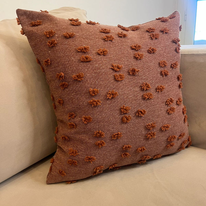 Rust textured square throw pillow with raised pom pom details placed on a beige sofa