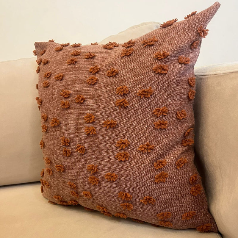 Rust textured square throw pillow with raised pom pom details placed on a beige sofa
