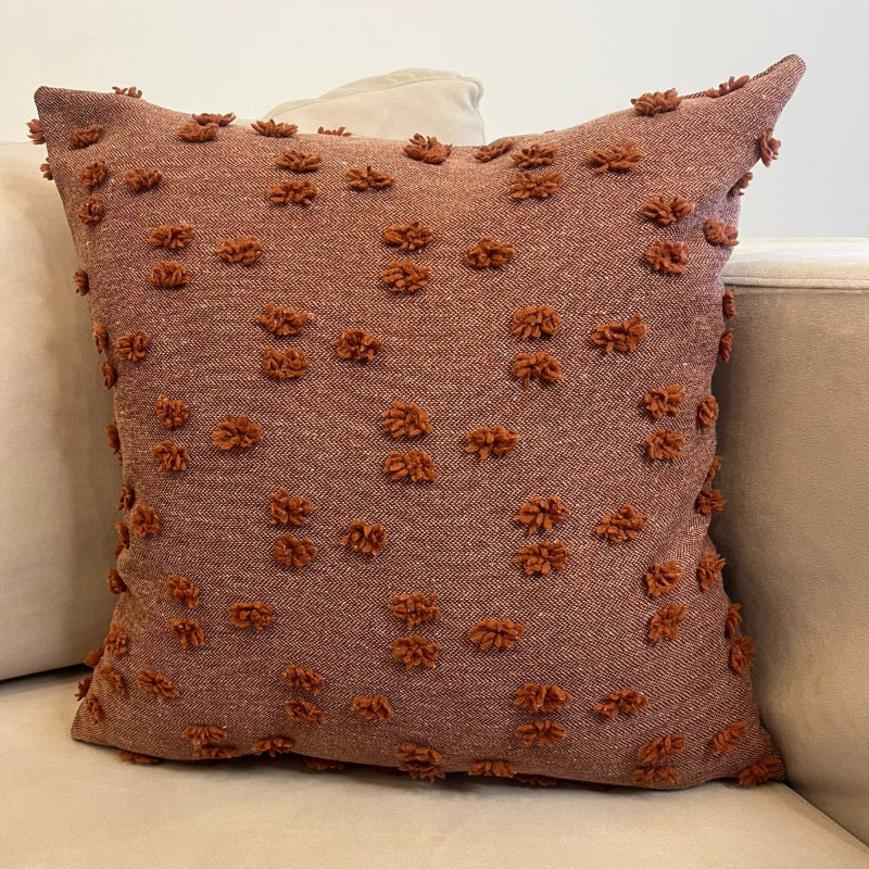 Rust textured square throw pillow with raised pom pom details placed on a beige sofa