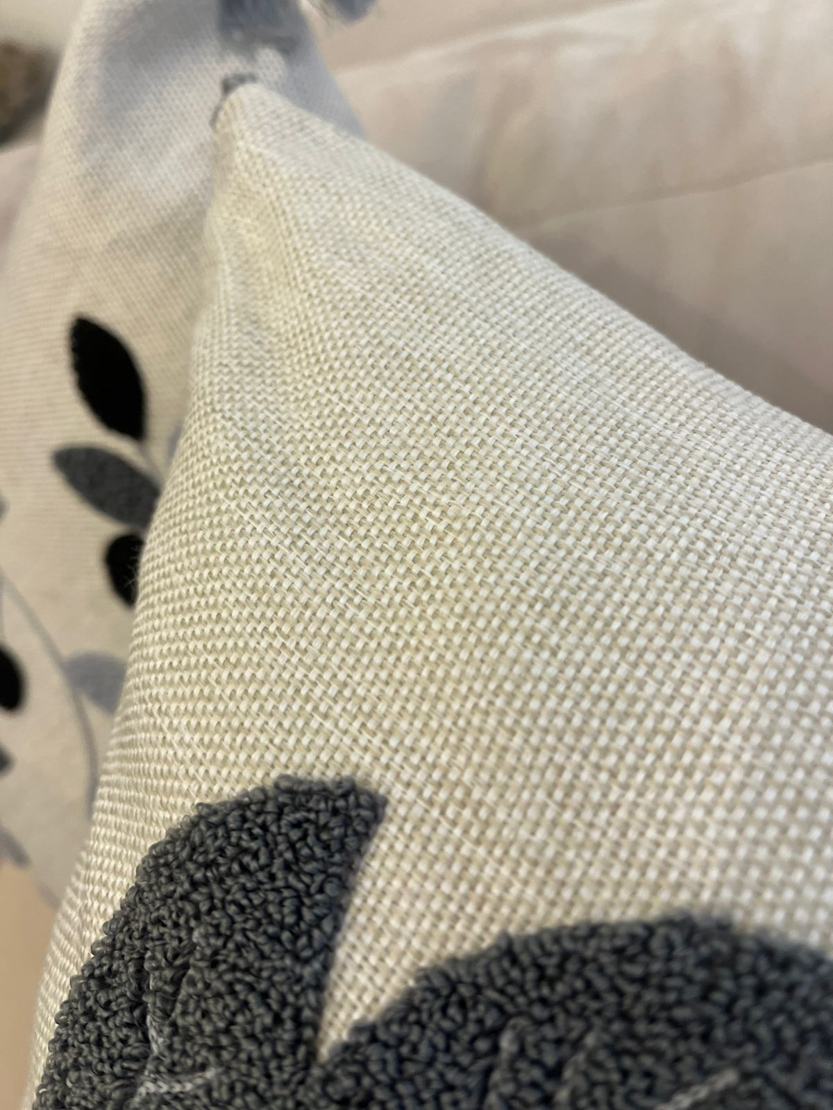 Square decorative pillow with embroidered grey and black leaf design and tassel corners placed on a beige sofa