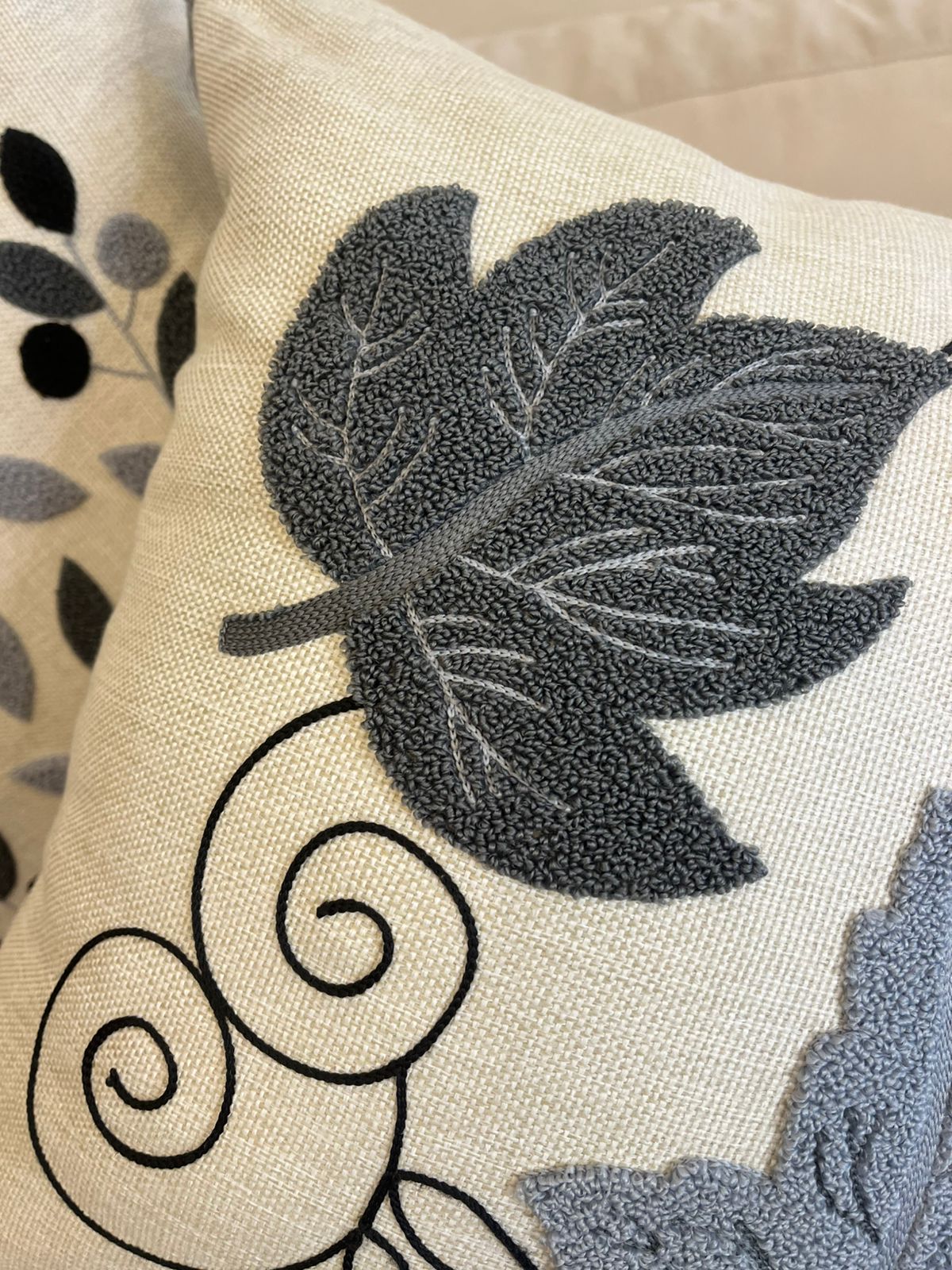 Square decorative pillow with embroidered grey and black leaf design and tassel corners placed on a beige sofa