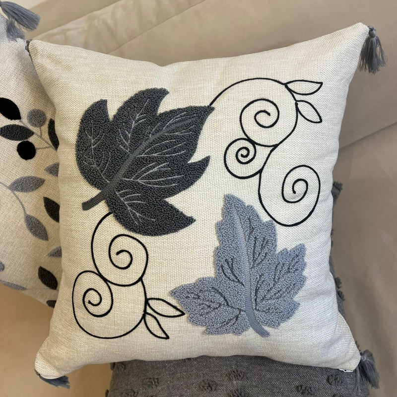 Square decorative pillow with embroidered grey and black leaf design and tassel corners placed on a beige sofa