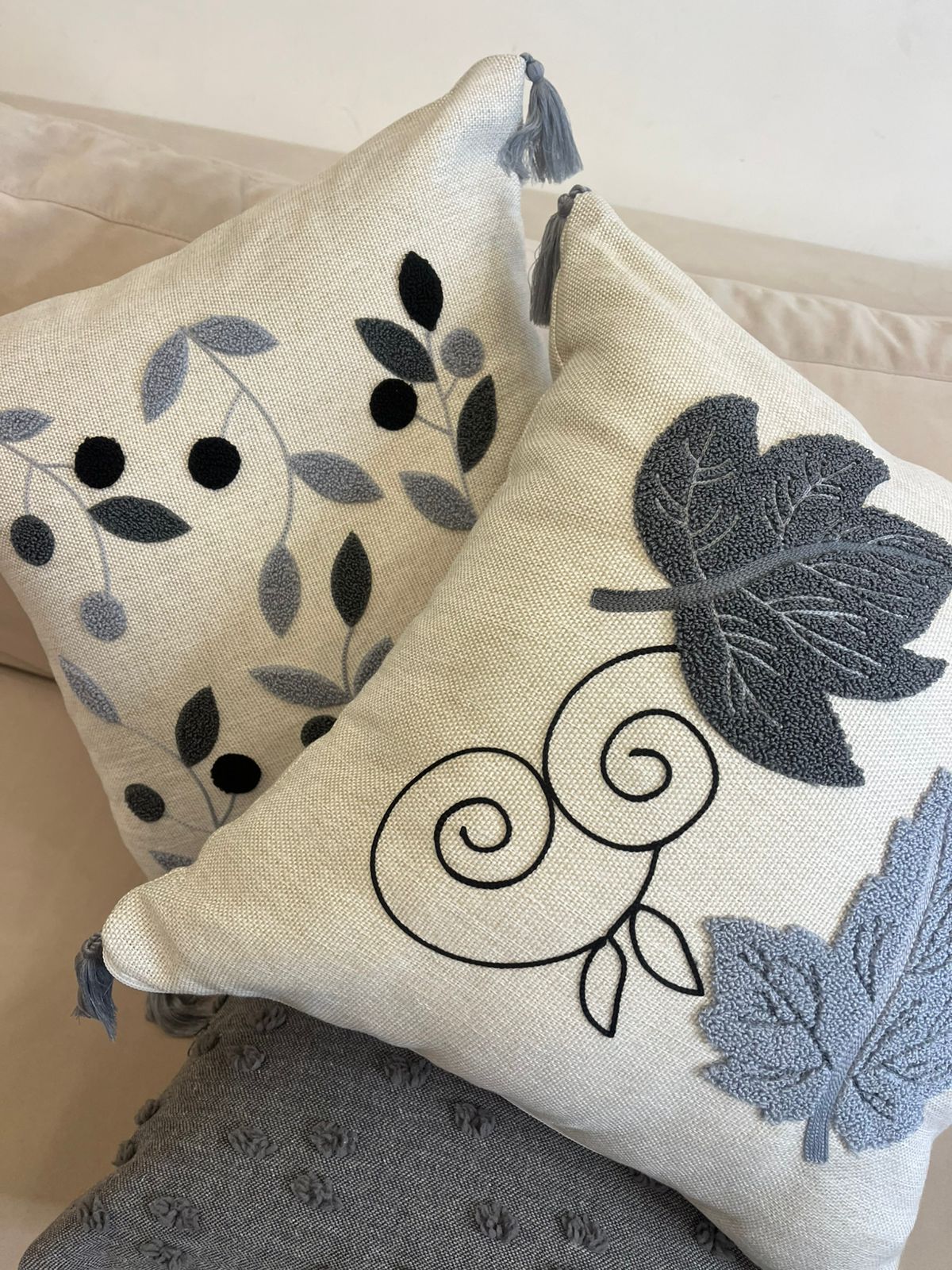 Square decorative pillow with embroidered grey and black leaf design and tassel corners placed on a beige sofa