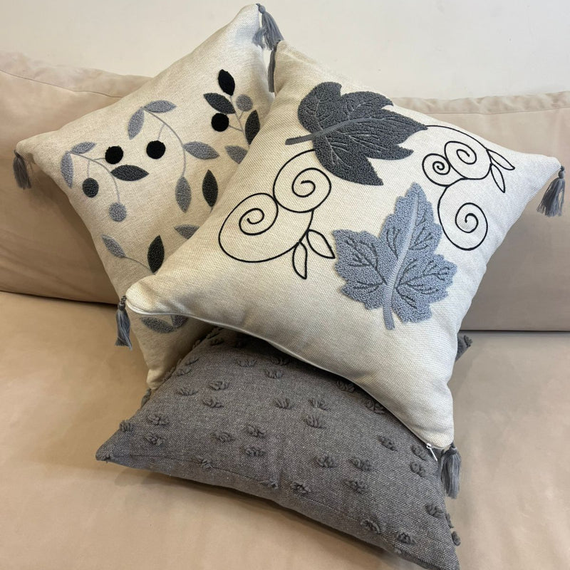 Square decorative pillow with embroidered grey and black leaf design and tassel corners placed on a beige sofa