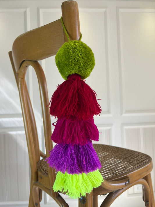 Green pom-pom tassel hanging decoration with red, magenta, purple, and neon green yarn tassels hanging on a wooden chair.