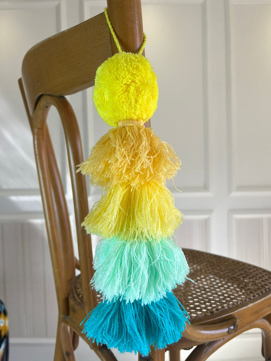 Yellow pom-pom tassel hanging decoration with layered yarn tassels in yellow, mint green, and turquoise hanging on a wooden chair.