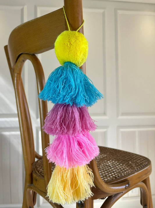 Handmade colorful pom pom tassel wall hanging with neon yellow pom pom and pastel pink, teal, purple, and yellow layered tassels on wooden chair