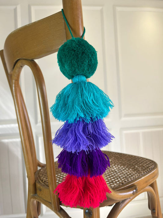 Handmade jewel-tone pom pom tassel hanging decor with layered teal, purple, and red yarn tassels displayed on wooden chair