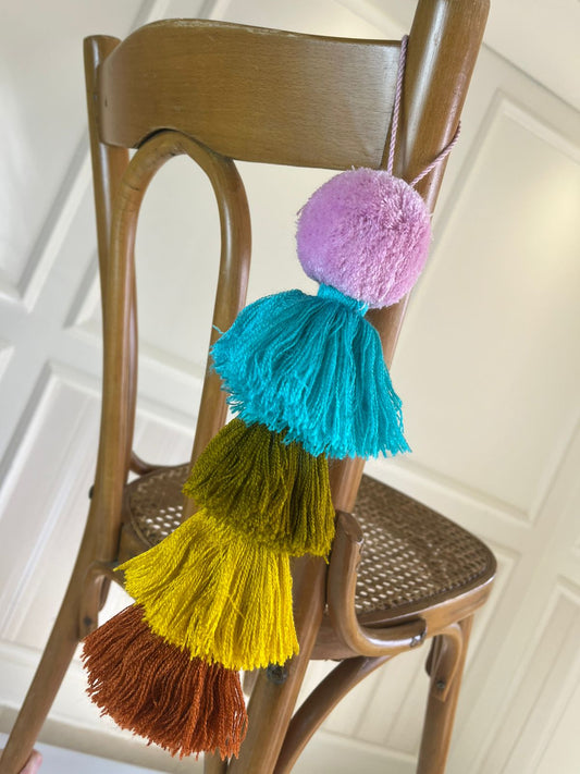 Handmade pom pom tassel hanging decor with teal, mustard, olive, and brown layered tassels displayed on wooden chair
