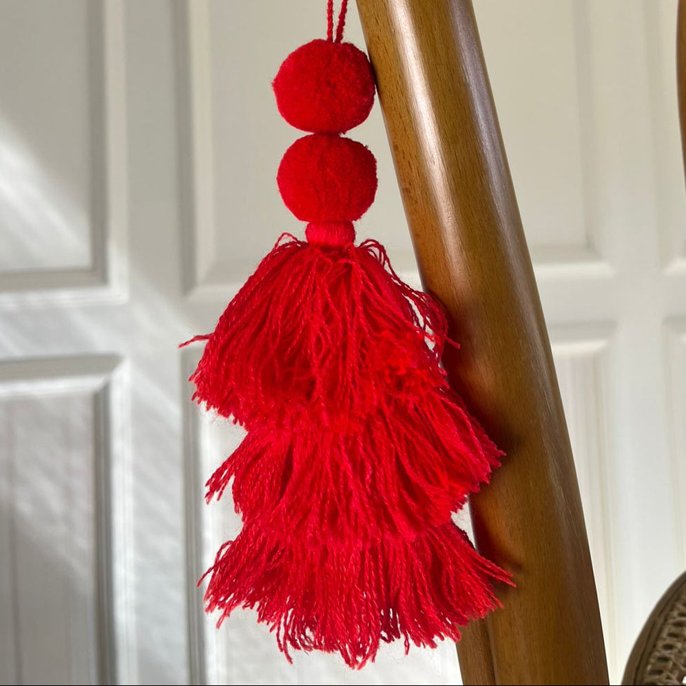 Classic red pom-pom tassel hanging decoration with two pom-poms and layered red yarn tassels hanging on a wooden chair.