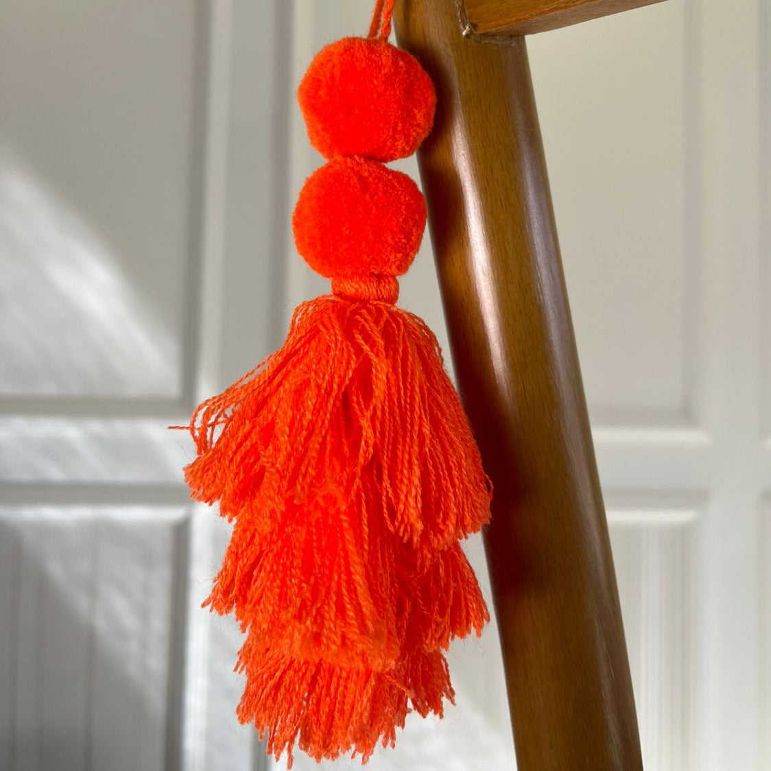 Neon orange pom-pom tassel hanging decoration with two pom-poms and layered neon orange yarn tassels hanging on a wooden chair.