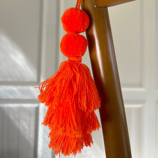 Neon orange pom-pom tassel hanging decoration with two pom-poms and layered neon orange yarn tassels hanging on a wooden chair.