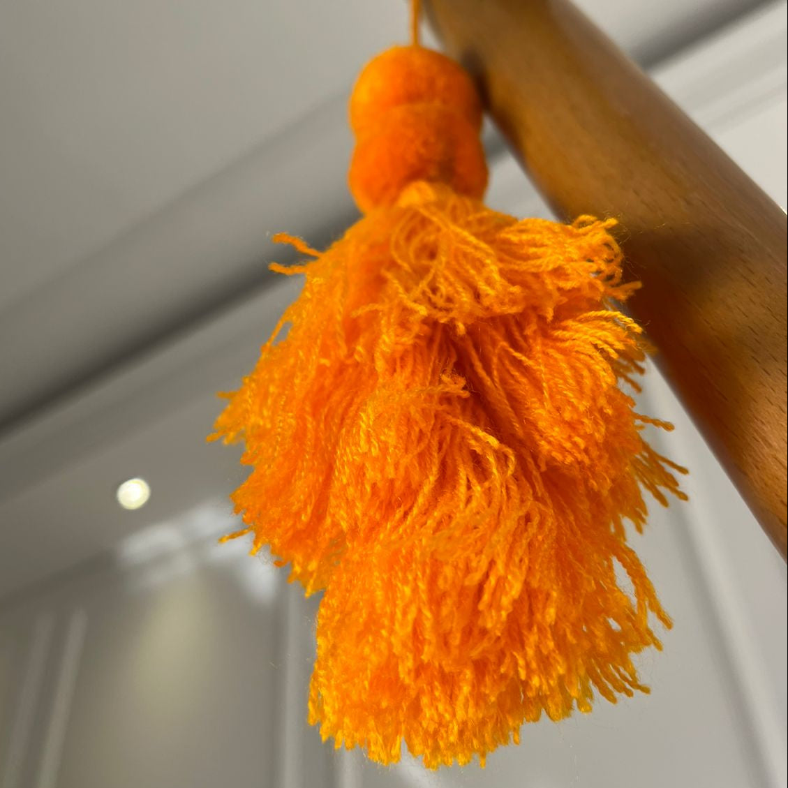 Orange pom-pom tassel hanging decoration with two pom-poms and layered orange yarn tassels hanging on a wooden chair.