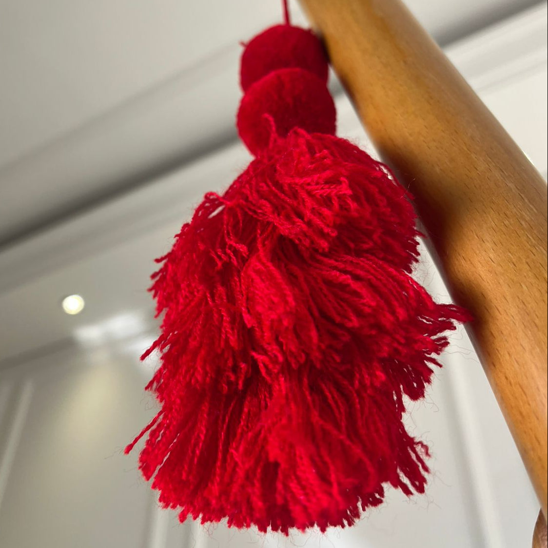 Red pom-pom tassel hanging decoration with two pom-poms and layered red yarn tassel hanging on a wooden chair.