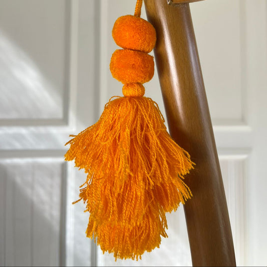 Orange pom-pom tassel hanging decoration with two pom-poms and layered orange yarn tassels hanging on a wooden chair.