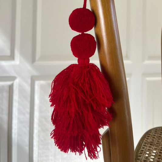 Red pom-pom tassel hanging decoration with two pom-poms and layered red yarn tassel hanging on a wooden chair.