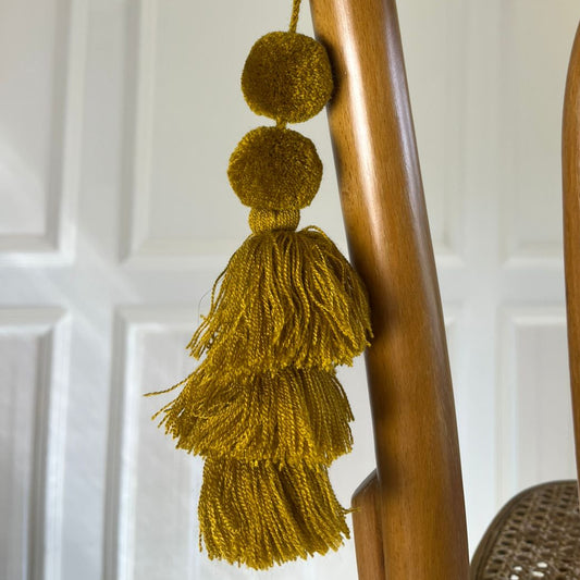 Mustard gold pom-pom tassel hanging decoration with layered yarn tassels hanging on a wooden chair.