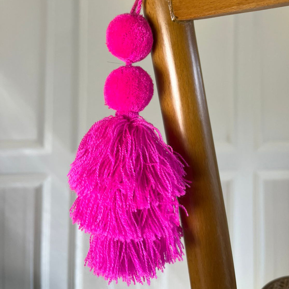 Hot pink pom-pom tassel hanging decoration with two pom-poms and layered pink yarn tassels hanging on a wooden chair.