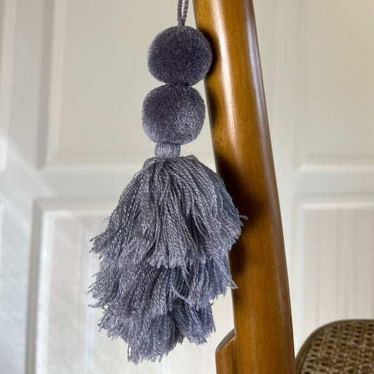 Grey pom-pom tassel hanging decoration with two pom-poms and layered grey yarn tassels hanging on a wooden chair.