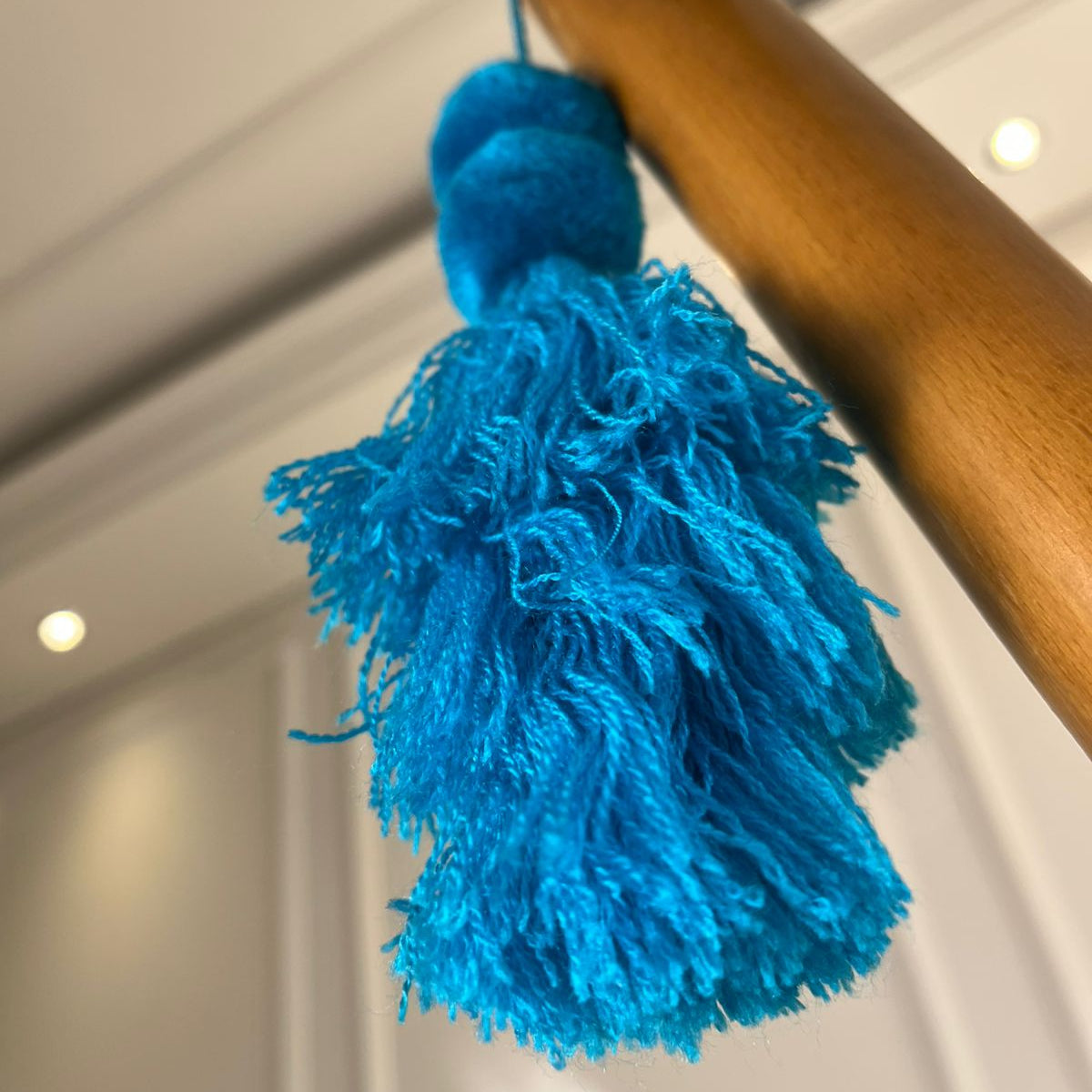 Turquoise blue pom-pom tassel hanging decoration with two pom-poms and layered turquoise yarn tassels hanging on a wooden chair.