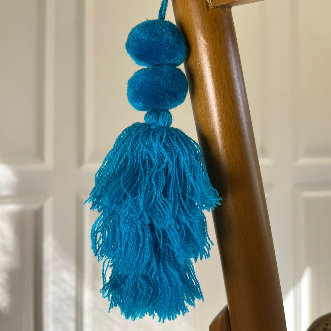 Turquoise blue pom-pom tassel hanging decoration with two pom-poms and layered turquoise yarn tassels hanging on a wooden chair.