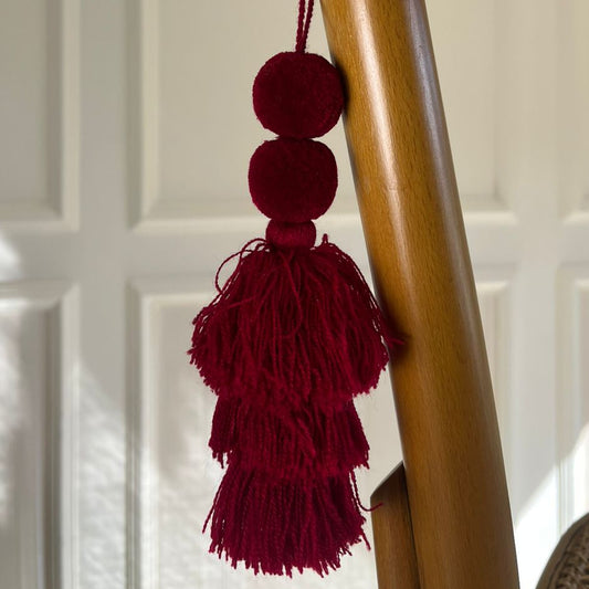Burgundy pom-pom tassel hanging decoration with two pom-poms and layered burgundy yarn tassels hanging on a wooden chair.