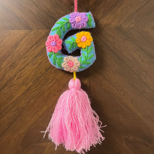 Handmade embroidered letter G ornament with colorful floral patterns and pink tassel on wooden background
