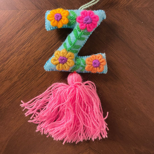 Handmade embroidered letter Z ornament with colorful floral stitching on blue fabric and pink tassel on wooden background