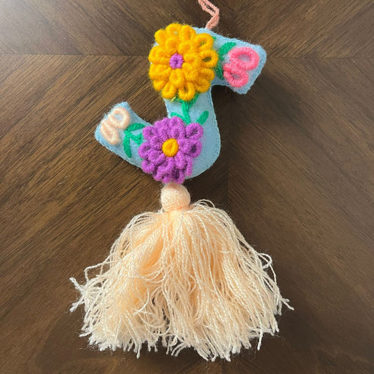 Handmade embroidered letter J ornament with colorful floral embroidery and peach tassel on wooden background