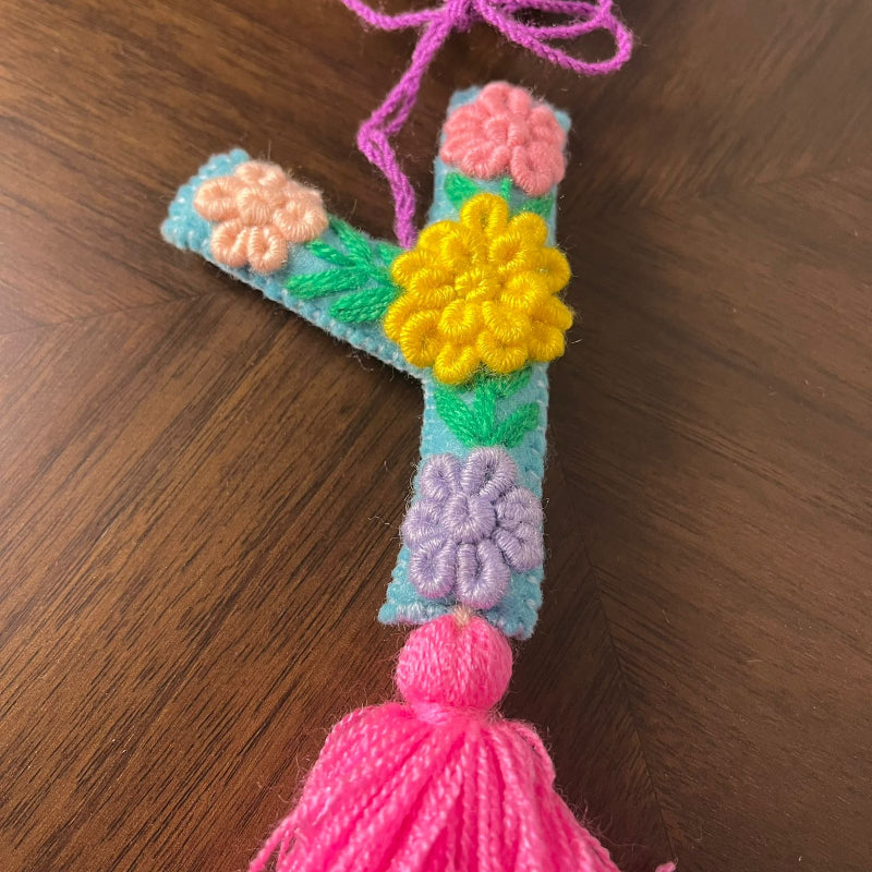 Handmade embroidered letter Y ornament with colorful floral stitching and bright pink tassel on wooden surface